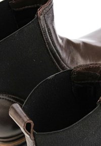 Gros plan de bottines Chelsea en cuir marron avec panneaux latéraux élastiques noirs et languettes de traction sur une surface claire.