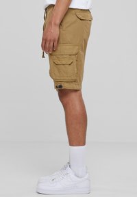 Beige cargoshorts met meerdere zakken, een taille met trekkoord en een gladde textuur. Gecombineerd met witte sneakers en enkelsokken.
