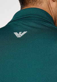 Krupan kadar prikaz osobe koja nosi košulju teksturirane tamnozelene boje s malim stiliziranim logotipom bijelog orla na gornjem dijelu leđa.