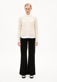 Crèmekleurig gebreid sweater met geribbelde zoom over een witte blouse met kraag, gecombineerd met hoogstaande, uitlopende zwarte ribfluwelen broek en zwarte schoenen.