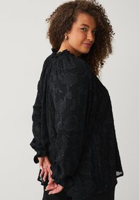 Sort bluse med blomstret struktur, lange ærmer, draperet udskæring og elastiske manchetter, i en løs pasform og blødt stof.
