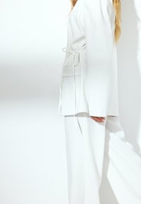 El traje blanco ajustado presenta una chaqueta entallada con un lazo en la cintura y pantalones de pierna ancha, confeccionado en una tela suave y ligera.