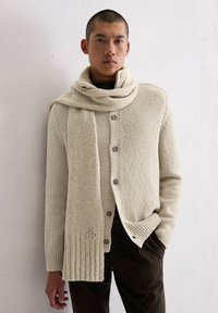 Cardigan in maglia beige con bottoni, abbinato a una sciarpa coordinata. Il tessuto ha una finitura strutturata. Logo visibile sulla sciarpa.
