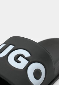 Sort gummisandal med en bred rem, der har indgraveret "HUGO" tekst i lyseblå. Glat overflade og afrundede kanter.