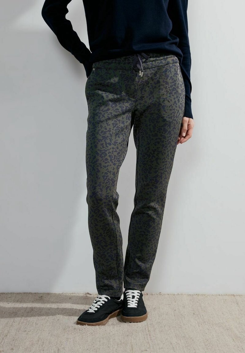 Personne portant un pantalon imprimé léopard noir et vert foncé, un pull noir et des baskets noires avec des lacets blancs, se tenant contre un mur clair.