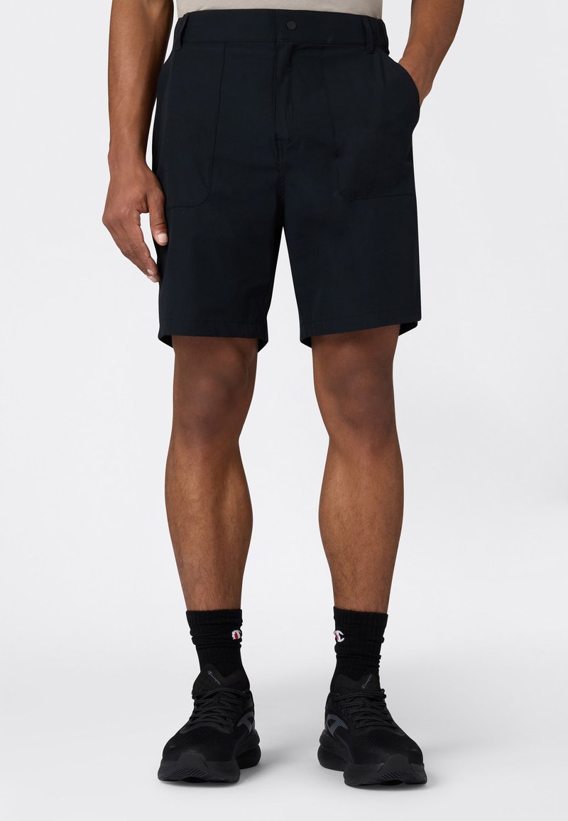 Czarne szorty z gładkiego materiału, z dwoma przednimi kieszeniami i luźnym krojem, zestawione z czarnymi butami sportowymi i skarpetami.