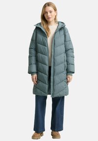 Gevulde tealjas met capuchon, met een ritssluiting aan de voorkant, chevron-gequilt ontwerp, zijzakken en ribgebreide manchetten, gedragen over een lichte trui en jeans.