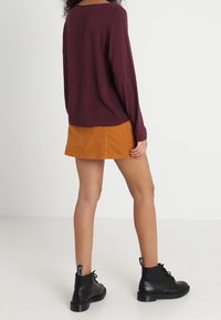 Maglia a maniche lunghe color mogano abbinata a una gonna gialla senape. Stivali neri alla caviglia con lacci e suola in gomma. Sfondo piatto.