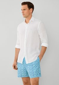 Muškarac nosi bijelu lanenu košulju s dugim rukavima, podignutim do lakta, i svijetloplave šortse s uzorkom, stoji s jednom rukom u džepu.