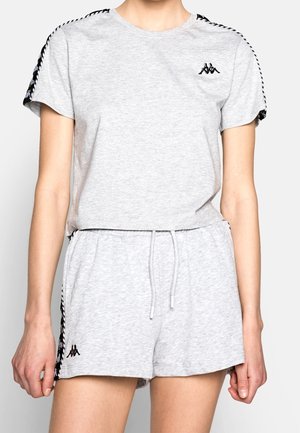 Kobieta ubrana w jasnoszary, krótko rękawowy t-shirt Kappa i pasujące szorty z czarnym logo i lamówką, stoi na tle jednolitego tła.
