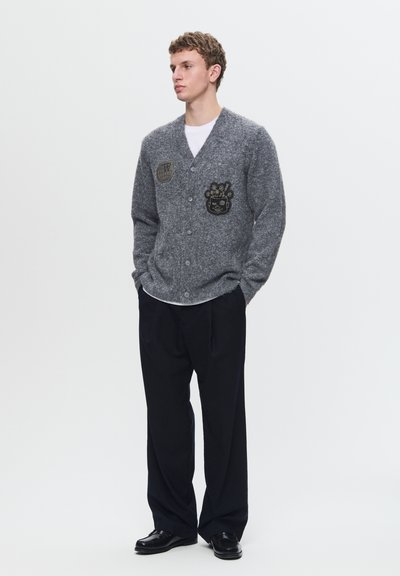 Grijze gebreide cardigan met V-hals, voorzien van twee geborduurde patches, knopen aan de voorkant en lange mouwen, gecombineerd met loszittende zwarte broek en schoenen.