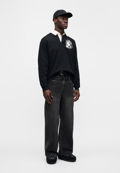 Μαύρο πόλο με μακριά μανίκια, εκρού γιακά και γραφικό αστροναύτη, συνδυασμένο με φαρδιά μαύρα τζιν παντελόνια και μαύρα πάνινα παπούτσια χωρίς κορδόνια.