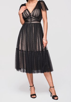 Frau trägt ein schwarzes, durchsichtiges Polka-Dot-Tüll-Midikleid mit flatternden Ärmeln und schwarzen High-Heel-Sandalen, stehend vor einem schlichten Hintergrund.
