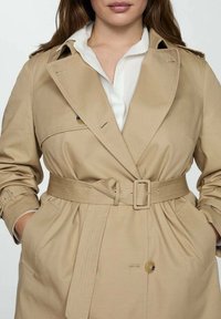 Trench-coat beige à taille ceinturée, à double boutonnage sur le devant et grandes poches. Confectionné dans un tissu lisse avec un col structuré et des accents de boutons.