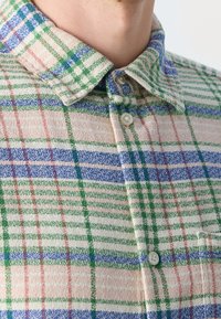 Koszula na guziki w kratę w kolorach zielonym, niebieskim, czerwonym i beżowym. Posiada kołnierzyk i teksturowaną tkaninę.