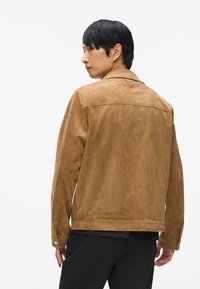 Rjava semiš jakna z gladko teksturo, s poudarjenim ovratnikom in vidnim hrbtnim šivom z detajlom.