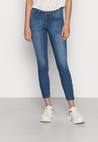 Smal passform blå denimjeans med medelhög midja, subtila slitningar, fem fickor och sydda detaljer, i kombination med beige sneakers.