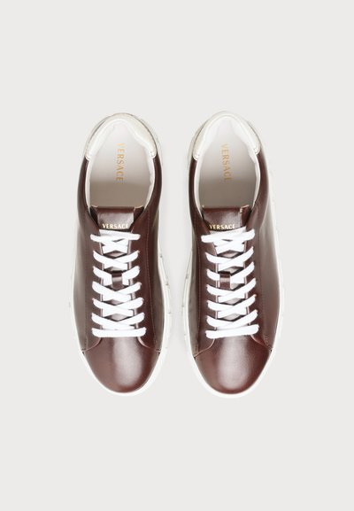Paire de baskets en cuir marron avec lacets et semelles blanches, marquage Versace sur la languette et la semelle intérieure, vue de dessus sur fond gris.