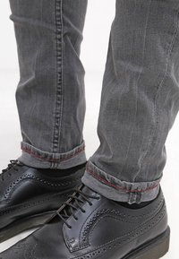 Zapatos de vestir de cuero negro con detalles de brogue, combinados con jeans de mezclilla gris que cuentan con un acento de costura roja en los puños.