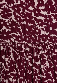 Burgundská látka s květinovým vzorem na krémovém pozadí; má na pasu nabrání, což vytváří texturovaný efekt.