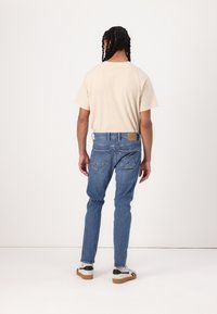 Tricou bej cu mânecă scurtă, asortat cu blugi albaștri din denim, având un eticheta texturată pe spate și un design cu cinci buzunare. Încălțămintea include adidași albi.