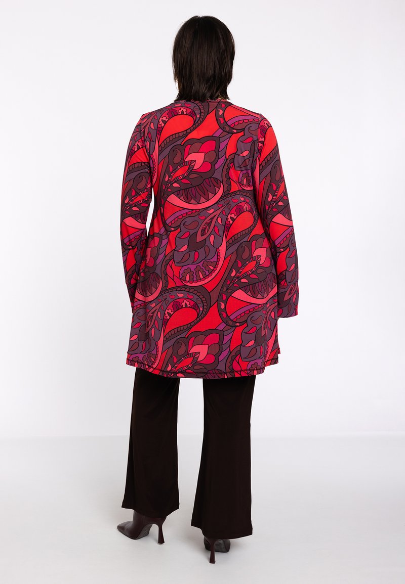 Tunique à manches longues avec un motif paisley rouge et violet audacieux, associée à un pantalon noir évasé et des chaussures à talons. Tissu lisse et coupe décontractée.