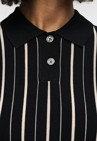 Polo noir avec fines rayures verticales beiges et blanches et deux boutons gris au col, porté par une personne à la peau foncée.