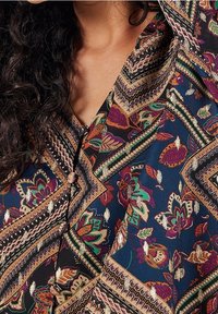 Blouse à motifs multicolores avec des motifs floraux en vert, violet et accents dorés sur un fond bleu marine. Col en V et boutons.