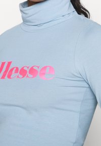 Helles blaues Rollkragenoberteil aus glattem Stoff, mit einem auffälligen pinken "ellesse"-Logo über der Brust, das ein figurbetontes Design betont.