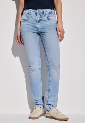 Light blue denim jeans with a slim fit, five-pocket design, silver button closure, and subtle fading. Paired with beige sneakers.