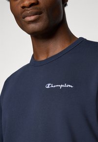 Marineblå bomulds t-shirt med rund hals, der har et hvidt broderet "Champion" logo på venstre bryst. Glat tekstur.