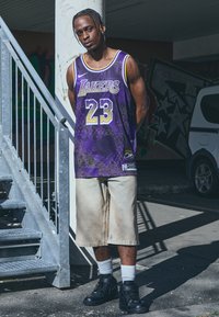 Maglia viola con accenti bianchi e dorati, con grafiche "Lakers" e "23". Pantaloni corti beige e sneakers nere completano l'outfit.
