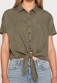 Camicia corta a maniche corte in cotone verde oliva con bottoni sul davanti e dettaglio a nodo sul fondo, indossata su un paio di jeans in denim blu chiaro.