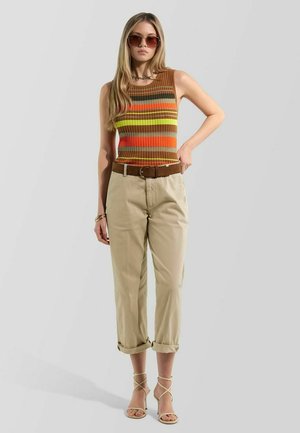 Mujer joven con gafas de sol, top sin mangas a rayas, pantalones beige remangados, cinturón marrón, pulseras doradas y sandalias de tacón con tiras, de pie.