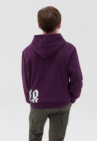 Sudadera morada con capucha, que presenta un diseño gráfico en blanco y menta en la parte posterior, combinada con pantalones cargo oliva.