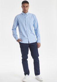 Camisa de botones de manga larga color azul claro con cuello, combinada con pantalones oscuros de corte navy y zapatillas blancas. La camisa tiene una textura suave.
