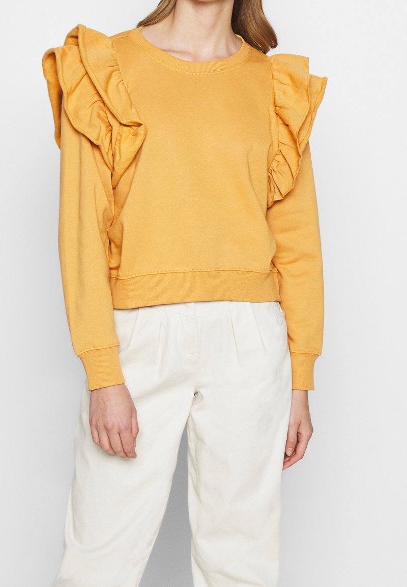 Sweat-shirt jaune avec des accents à volants sur les épaules, tissu doux, manches longues et ourlet côtelé. Porté avec un pantalon de couleur claire.