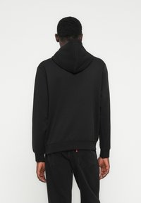 Άνδρας που φοράει μαύρο φούτερ με κουκούλα και μαύρο τζιν, στέκεται με την πλάτη στην κάμερα, μπροστά σε απλό λευκό φόντο.