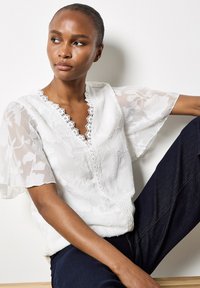 Weiße Bluse mit floralem Muster und transparenten Ärmeln, Spitzenbesatz am Ausschnitt und Knopfdetails auf der Vorderseite, kombiniert mit dunkelblauen Jeans.