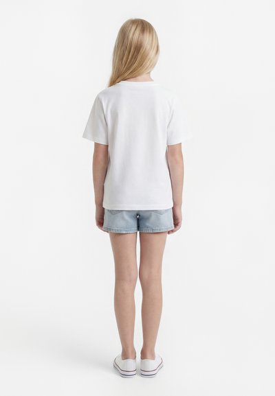 Jeune personne aux longs cheveux blonds portant un t-shirt blanc uni, un short en jean clair et des baskets blanches, debout de dos.