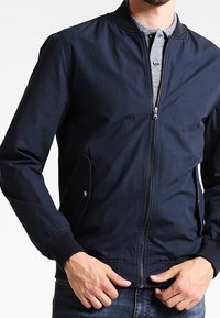 Marinblå bomberjacka med dragkedja, ribbade ärmslut och sidofickor. Slät textur, avslappnad design, bärs över en grå pikétröja.