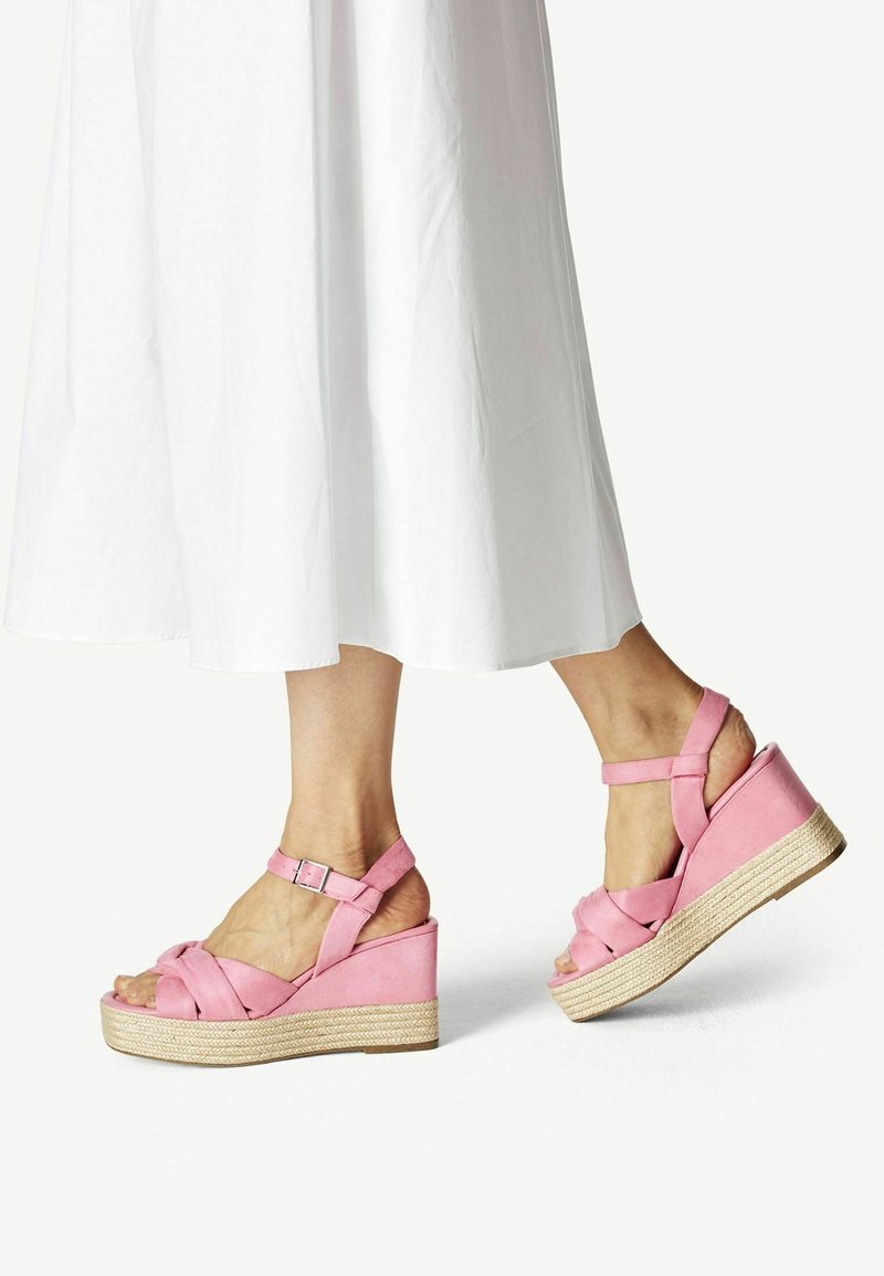 Sandales compensées en daim rose avec design de brides croisées, fermeture à boucle et plateforme enveloppée de jute. Associées à une robe blanche longueur cheville.