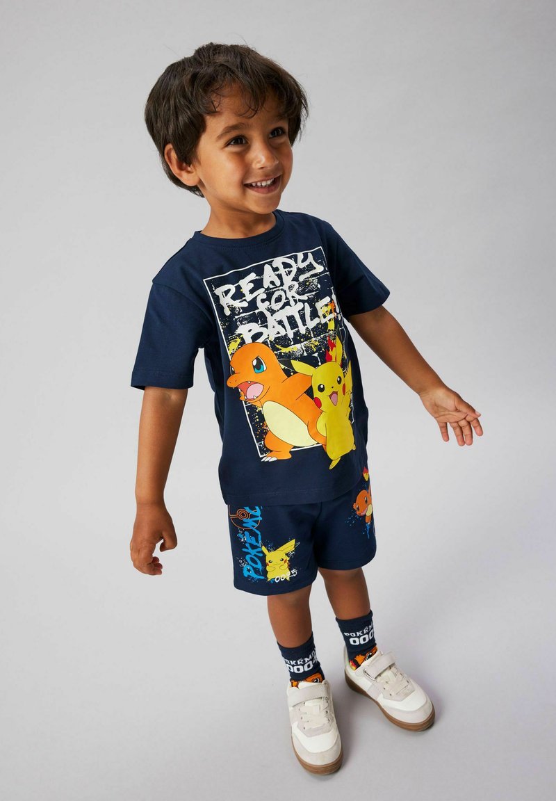 Garçon souriant portant une tenue Pokémon bleu marine avec des motifs Pikachu et Charmander, des baskets blanches et des chaussettes à thème Pokémon.