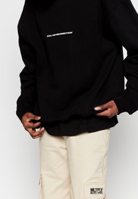 Sudadera negra con el texto bordado en blanco "ALMA Y RESURRECCIÓN" en la parte delantera, llevada sobre pantalones cargo beiges con un acento de bolsillo.