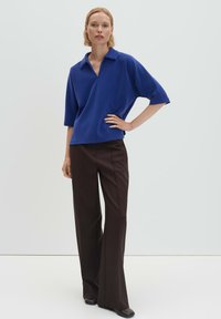 Femme portant un haut bleu à col montant et manches mi-longues, avec un pantalon large taille haute de couleur marron foncé, debout avec une main sur la hanche.