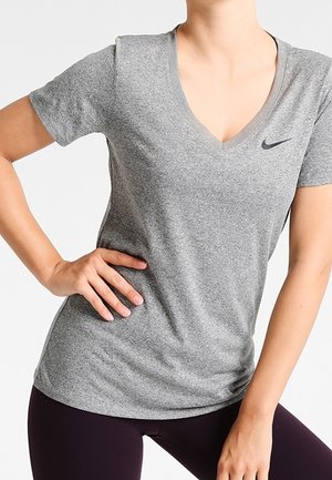 Osoba ubrana w jasnoszarą koszulkę Nike z dekoltem w serek i ciemne legginsy, z jedną ręką na biodrze i lekko obróconym tułowiem.