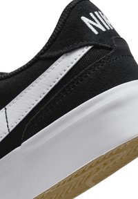 Fekete sportcipő texturált felsőrésszel és fehér oldalcsíkkal. Fehér gumitalp jellegzetes profilmintázattal. Nike logó a saroknál.