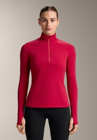 Top aderente a maniche lunghe rosso con collo alto e chiusura a mezza zip. Texture liscia con fori per i pollici nelle maniche, abbinato a leggings neri.