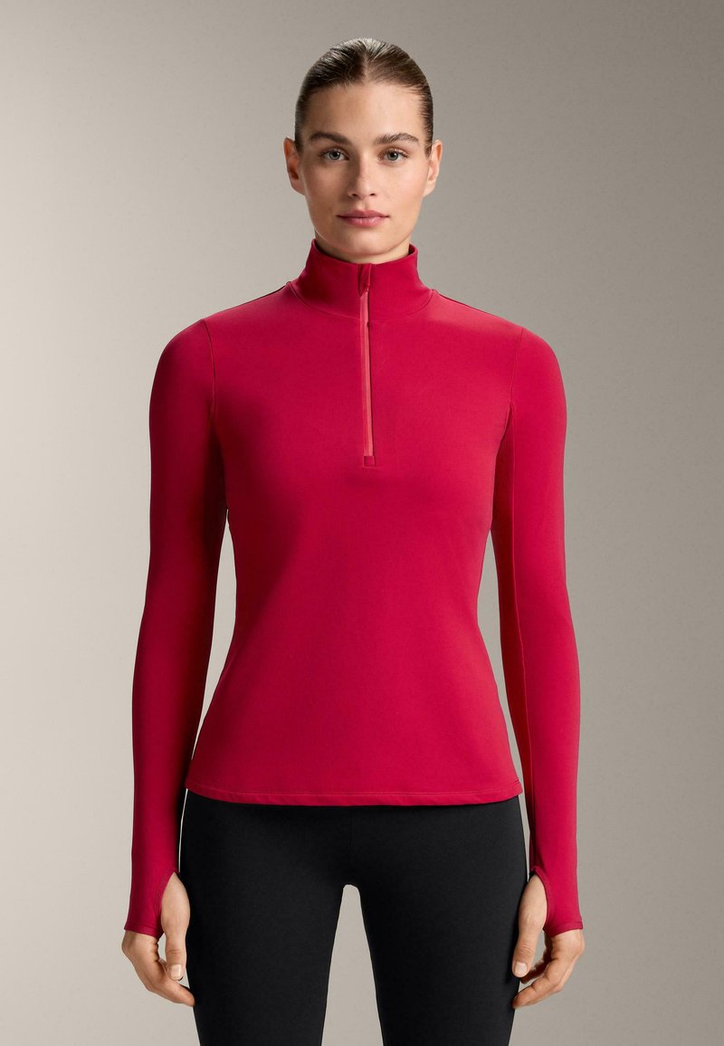 Top aderente a maniche lunghe rosso con collo alto e chiusura a mezza zip. Texture liscia con fori per i pollici nelle maniche, abbinato a leggings neri.