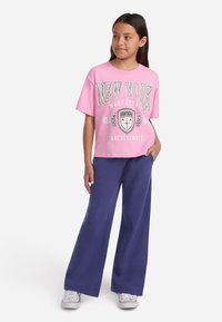 Ružová bavlnená tričko s potlačou "NEW YORK" a bielym písmom, spárované s modrými nohavicami so širokými nohavicami. Obsahuje biele tenisky.